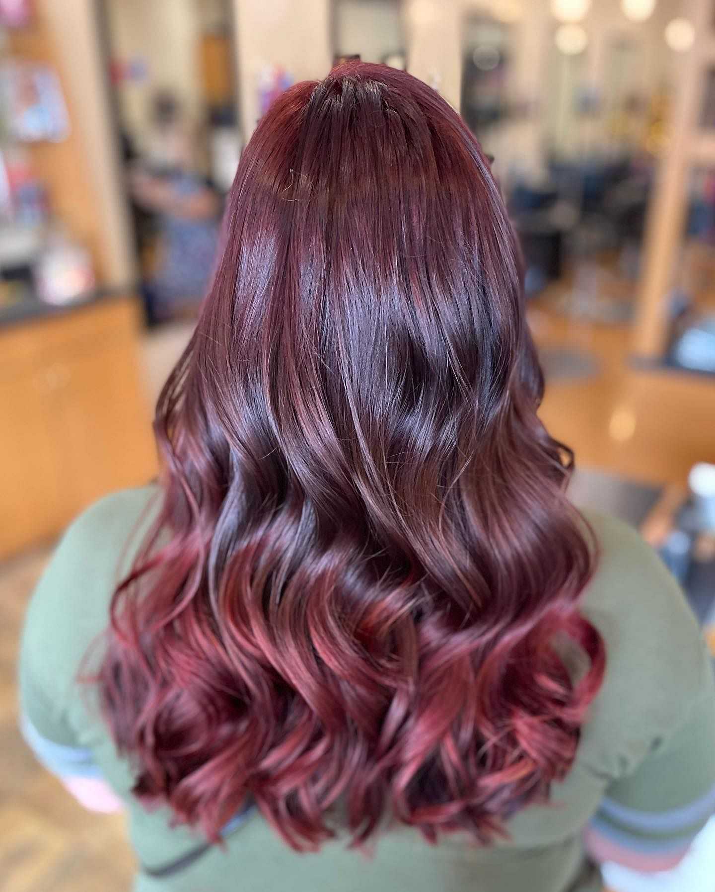 Woman with long, wavy brown hair with red highlights in a salon, viewed from behind.